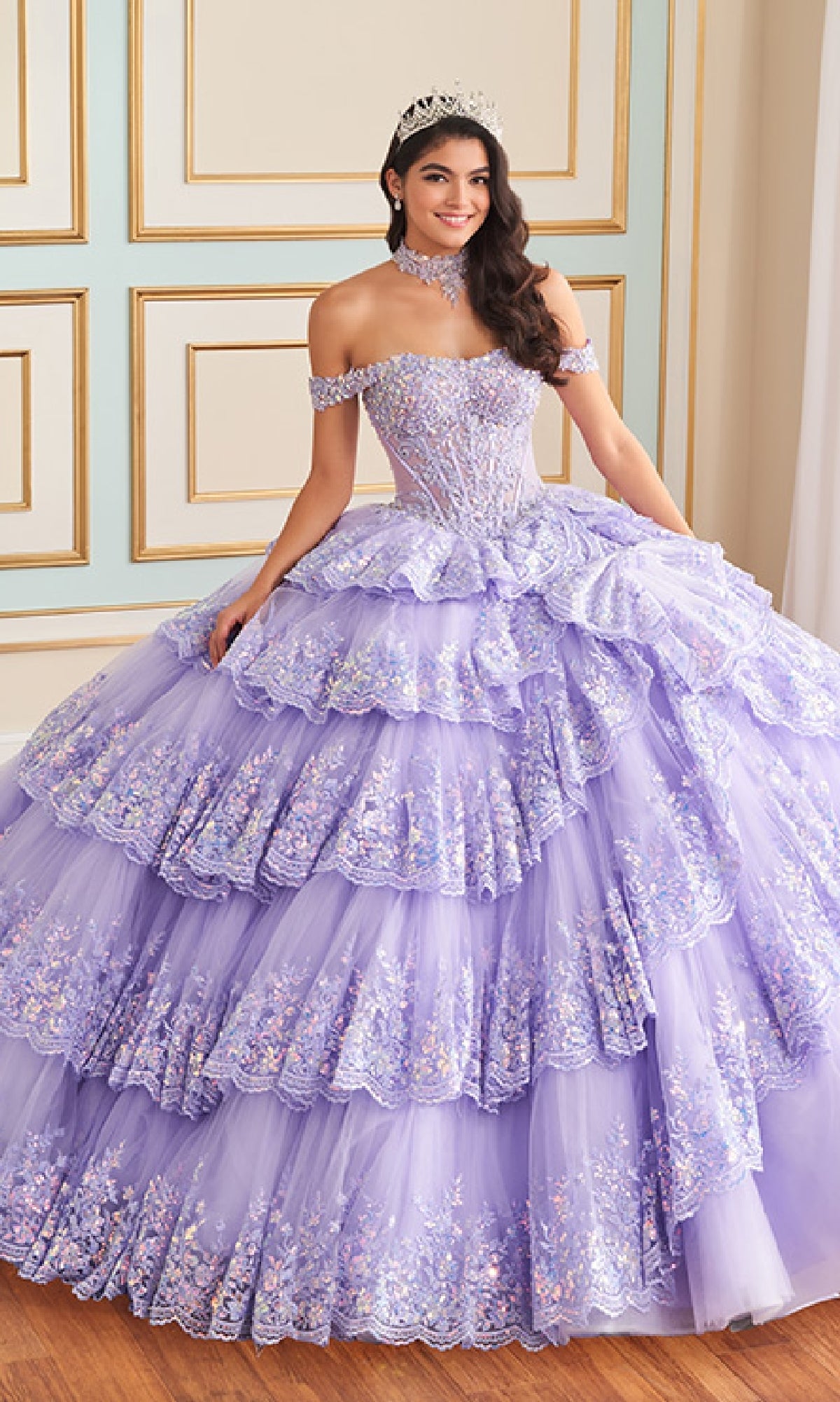 A young woman stands indoors against a decorated wall, wearing the Princesa Quinceanera Dress PR30181 in lavender with layered ruffles, floral embellishments, and a tiara.